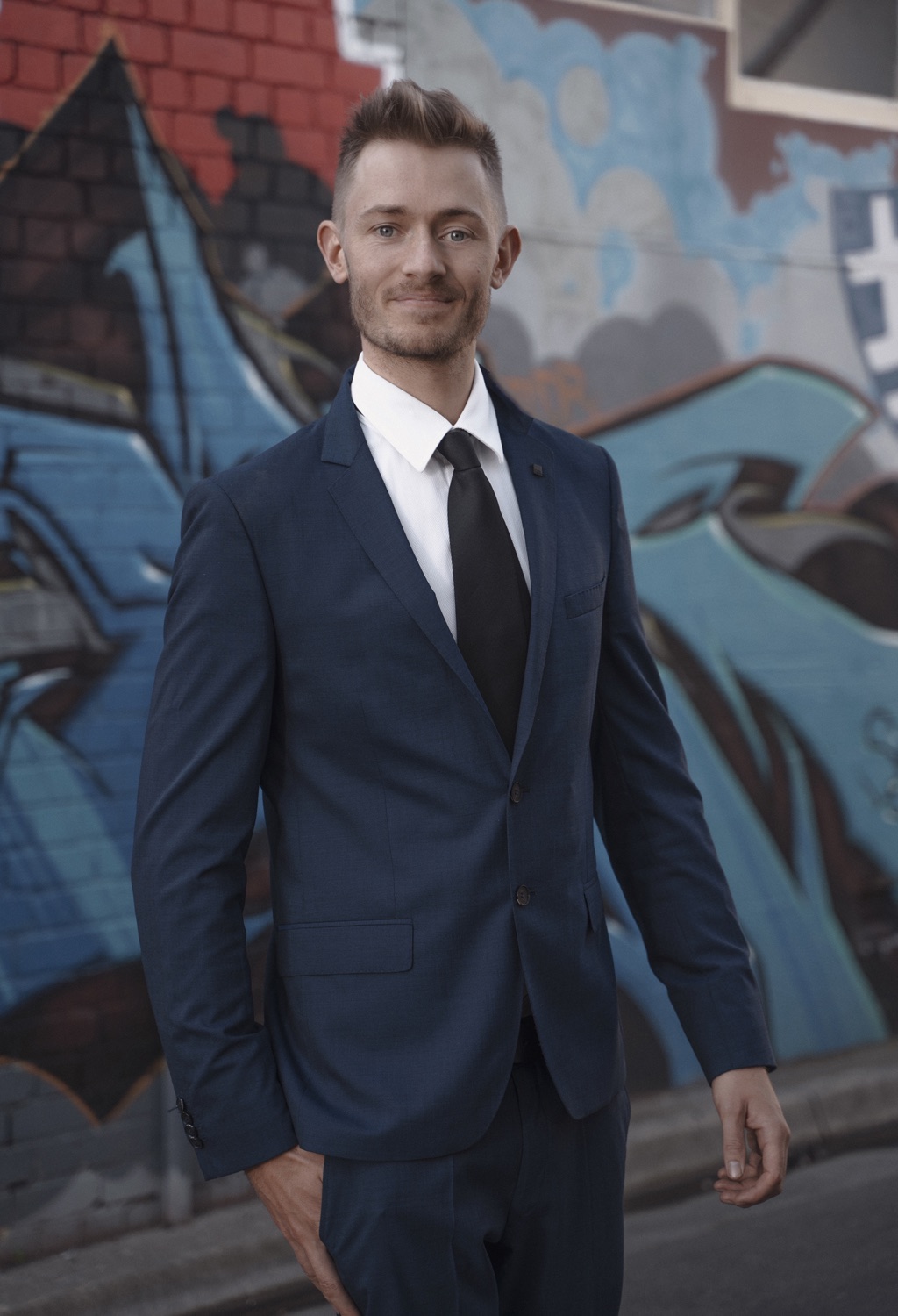 Andy Campbell, mindset coach, standing in a navy suit in front of a painted laneway wall